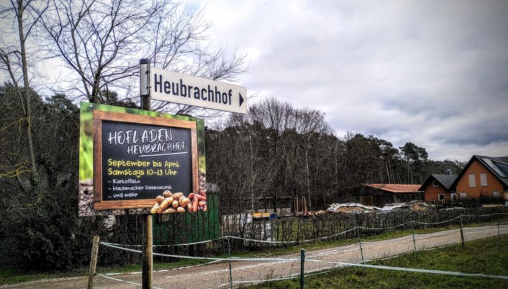 Hinweisschild des Hofladen Heubrachhof in Kleinostheim, der Bauernhof im Hintergrund.
